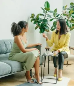 african american woman and asian american woman sitting down chatting with a large plant behind them